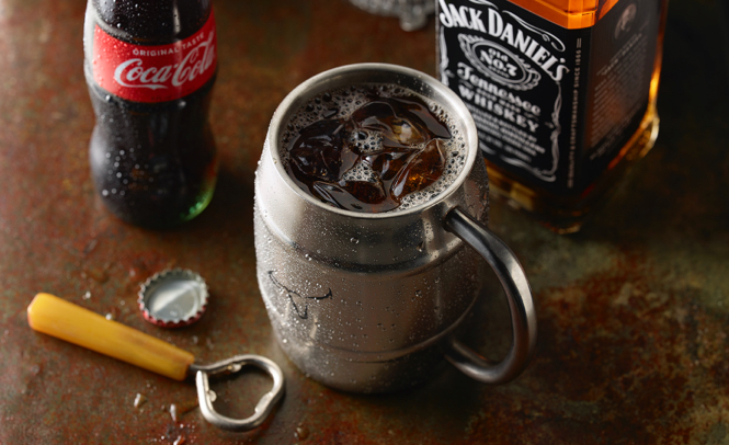 A metal mug containing a cocktail with a bottle of Coca Cola and Jack Daniels behind it