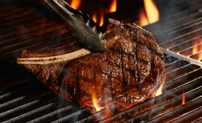 Tongs placing a bone-in steak on a flaming grill